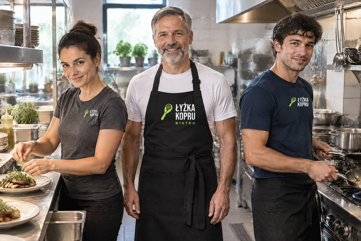 Wizualizacja AI: zespół fikcyjnego bistro Łyżka Kopru w kuchni — kucharka w grafitowej koszulce damskiej JHK TSRL CMF, kucharz w białej koszulce MALFINI Classic New i czarnym fartuchu z napierśnikiem JHK WKPARIS oraz kucharz w granatowej koszulce MALFINI Classic New i czarnej zapasce; przykładowe logo bistro na piersi koszulek i duże na napierśniku fartucha.